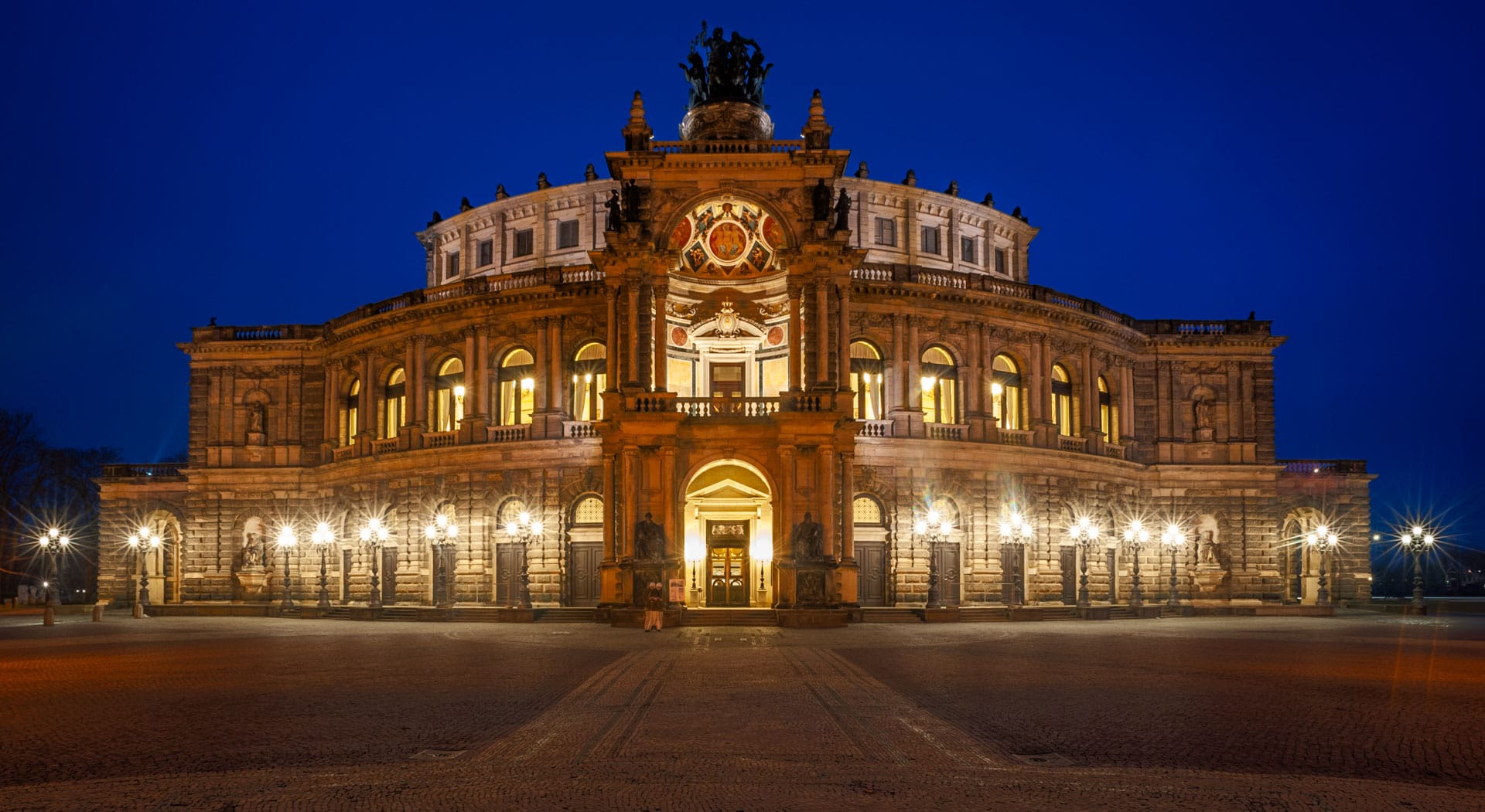 The illuminated facade of the Semperoper in Dresden against a deep blue evening sky, the wet pavement of the square reflecting the light